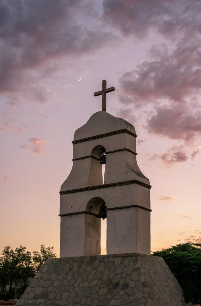 Silhouette of a Spanish mission bell tower with crescent moon and pink sky in California.